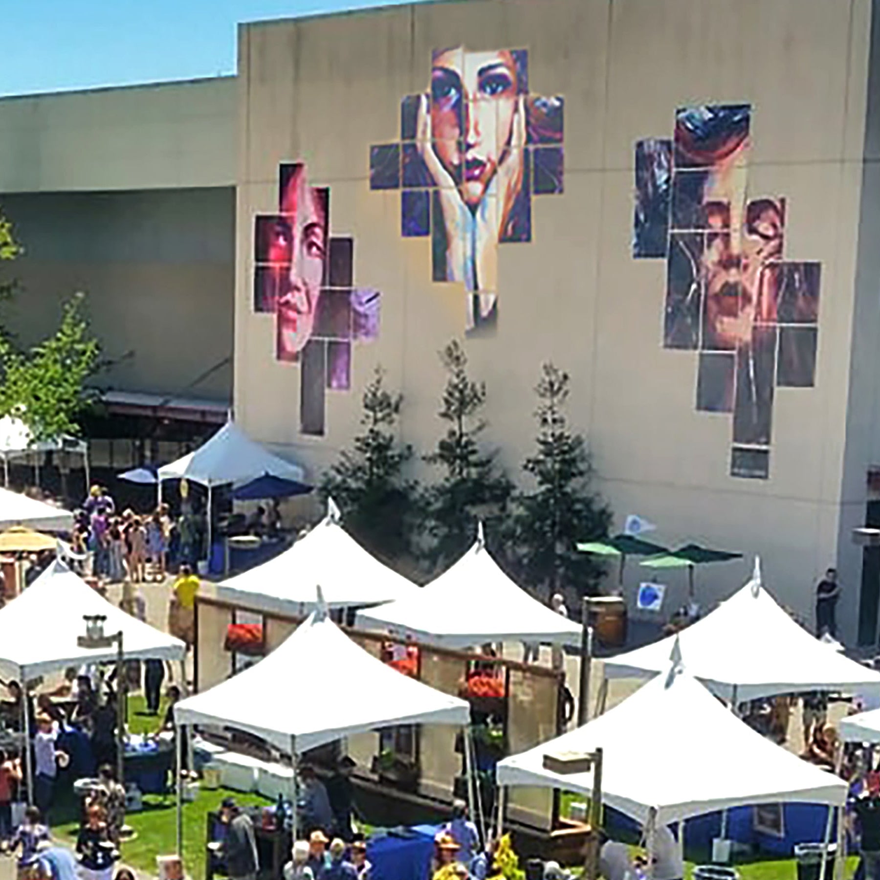 Outdoor event with tents and people in front of a building with a large mural.